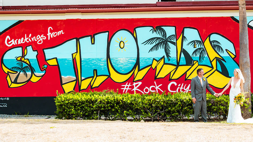 Husband holding wife's hand in front of Greetings from St Thomas mural in the USVI.